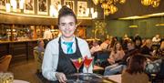 A photo of a waitress holding a tray of cocktails in a busy bar area.