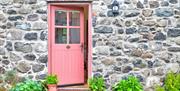 A pink wooden door of a converted cobblestone barn on a deck with plants.