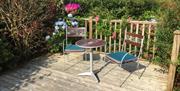 A decked outside seating area for two with bushes and flowers.