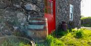 Front view of an old stone cottage with a red front door, and a wooden barrel and some metal vintage items along the front