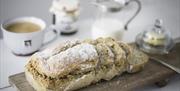 Image is of freshly baked bread and butter on a wooden tray with cup of tea, jam and milk in the background