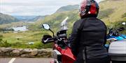 Image shows cyclist sitting on motorcycle enjoying a scenic view of countryside and lake