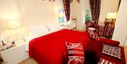 A double bedroom with a red bed cover and cushions, red  footrests, a red desk and a window.