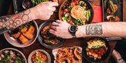People with tattooed arms reaching over a table for tapas food.