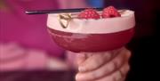 A close-up of a vibrant French Martini cocktail in a coupe glass, garnished with fresh raspberries and a decorative skewer. A person in a pink top hol