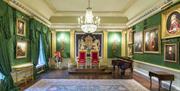 Image is of a room with wooden floor, large rug, various paintings on the wall and 2 throne chairs