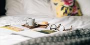 Coffee and traybake on a tray along with a magazine and a pair of eye glasses on a bed