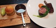 Image is of plate with type of meat on it and separate sauce in small saucepan with potato sitting beside it on a wooden serving board