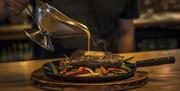 sizzling steak platter on table with sauce being poured over the steak from a gravy jug.