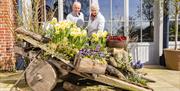 A man and a woman are standing behind a garden installation. The installation is an old cart which as boxes of flowers standing on it, including a dis