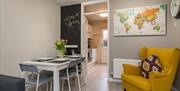 A photo of an area with a dining table, a world map, a, yellow chair and a doorway to a kitchen.