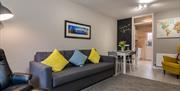 A photo of a living room with a sofa and chair which leads to a dining table and kitchen entrance.