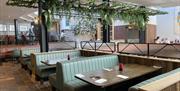 The Yard Restaurant, green booth seating with wooden tables. Green foliage hanging above from ceiling frame.