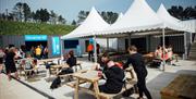 Image shows outside seating area with people eating and drinking at picnic tables