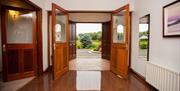 Image is of hallway with 2 sets of double doors opening out to show view of driveway and shrubbery