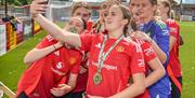 a girls football team pose for a selfie with their medals wearing red Manchester United uniforms