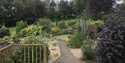 A paved path through a well planted garden with a bench leading to a forested path.