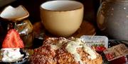 Close-up of a homemade scone topped with butter and dusted with powdered sugar, accompanied by a fresh strawberry and whipped cream. A ceramic teapot,