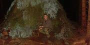 A man in camouflage clothes under a shelter made from trees.