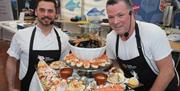 Two chefs wearing black aprons showing a table full of local seafood