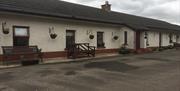 Image shows outside of cottage. Wooden benches outside. Gravel driveway.