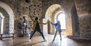 A woman and child having a pretend sword fight in the room at the top of the castle keep with the sun beaming through the window behind.