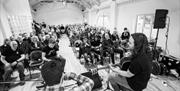 Black and white image of 2 men facing an audience.  One man is playing the violin and the other is playing a guitar.  The room is full and there