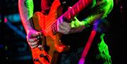 Close up image of an electric guitar being played. The guitarist has a tattoo on his arms and is wearing dark jean and a dark coloured t shirt.