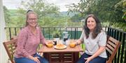 2 young women sitting at a patio table on balcony with tress in the background