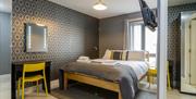 A double bedroom with grey wallpaper, a desk with a yellow chair, a mirror and a TV.