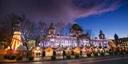 Belfast City Hall light up at night with Christmas market stalls in foreground in front of the City hall front gates.