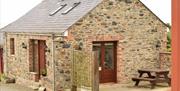 Image shows front and side of cottage. Wooden doors, gravel driveway and concrete patio with picnic table and parking area.