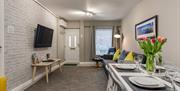 A photo of a living room with a TV, seats, a dining table and the houses front door.