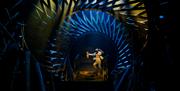 Actor Conor Quinn dressed as a teenager in shorts and a fairisle jumper gallops down some golden stairs on stage in the middle of a set that resembles