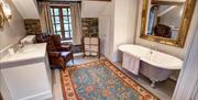 Image shows large bathroom with roll top bath and large mirror above it. Large rug. armchair, wash basin with storage underneath