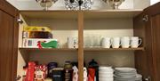 Wooden cupboard filled with glasses, mugs, plates, bowls tea, coffee and cereals.