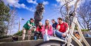 Steven pours a cup of tea for four tour guests sitting underneath the giant bronze sculpture of Aslan at CS Lewis square, in the foreground is the whe