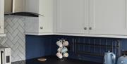 Grey wall and base cupboards, stainless steel cooker, microwave and extractor fan, white under-counter fridge and blue kettle and toaster.
