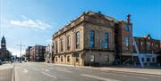 The Corn Exchange, a large light-coloured stone building with arched windows, stands on a quiet city street under a clear blue sky. There are addition