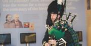 A man in traditional Scottish attire - green and blue tartan - plays the bagpipes. He is standing in front of an informative display about Robert Burn