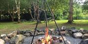 A fire pit with a hanging grill tray in a forest with picnic tables.