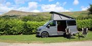 Image of a grey Volkswagen campervan parked on a grass patch in Co. Down. Camping chairs and a table is positioned outside the campervan.