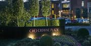 Image of the outside of the Crowne Plaza Belfast hotel. Flower bed with "Crowne Plaza" sign in the foreground, hotel and parking lot in the background