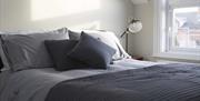 A double bed with cushions and a bedside table with a lamp beside a window.