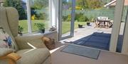 Conservatory sitting area with a chair leading to a sunny patio and garden with patio furniture.