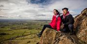 A couple sitting down on the edge of Slemish with surrounding countryside in view