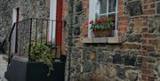 Image show front of stone property with flowerbed on window sill and wrought iron gate into red doorway