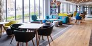 Photo of the open lobby in Holiday Inn Belfast City Centre. There are tables and chairs, a sofa with coffee tables and large bay windows bringing natu