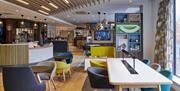 Photo of the open lobby in Holiday Inn Belfast City Centre. There are tables and colourful chairs. The bar and cafe is on the left hand side.
