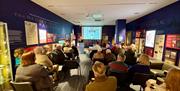 A group of people sit in rows of two or three in a warmly lit room - the Montgomery Gallery inside the Discover Ulster-Scots Centre. They are facing a